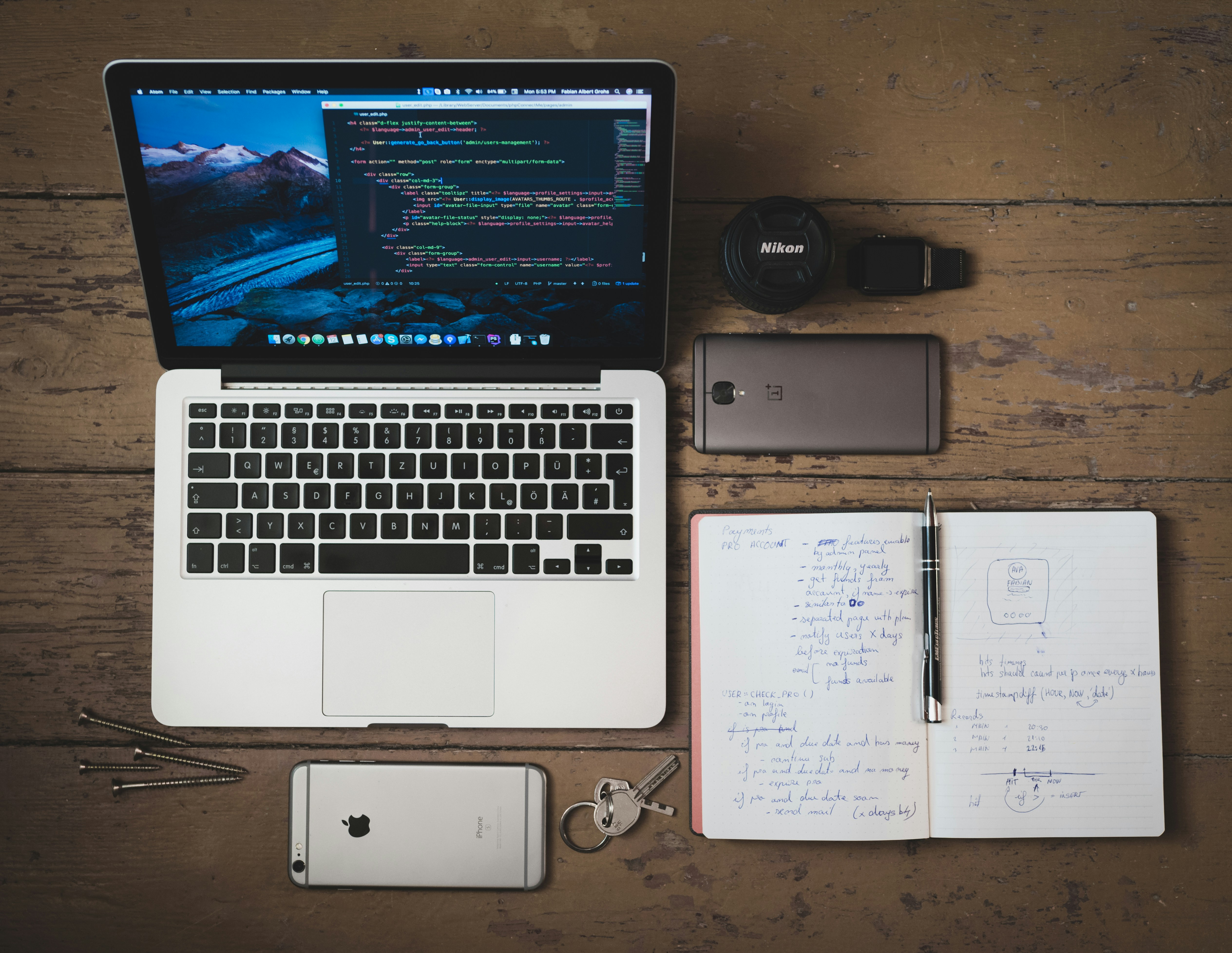 Overhead view of a laptop displaying code, smartphone, notebook, keys, and camera gear arranged on a wooden desk.