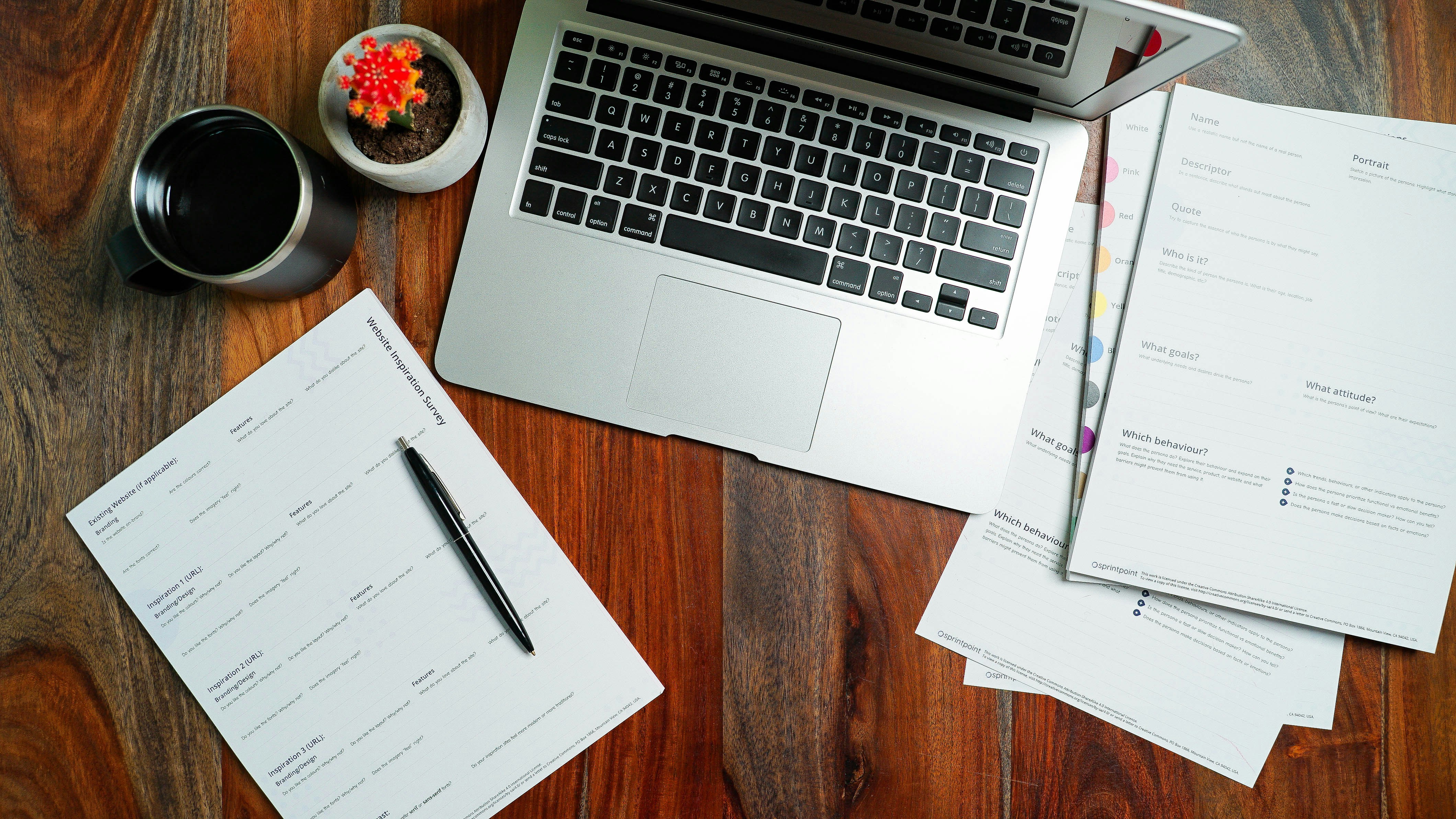 Overhead view of a laptop, printed planning worksheets, pen, coffee mug, and small plant on a wooden desk.