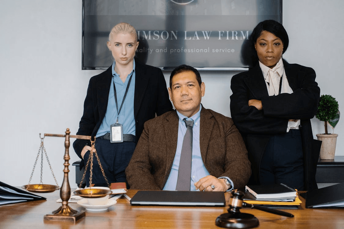 Three lawyers posing in a law office - one seated behind a desk, two standing behind him—with a gavel and balance scales on the table and a firm sign in the background.