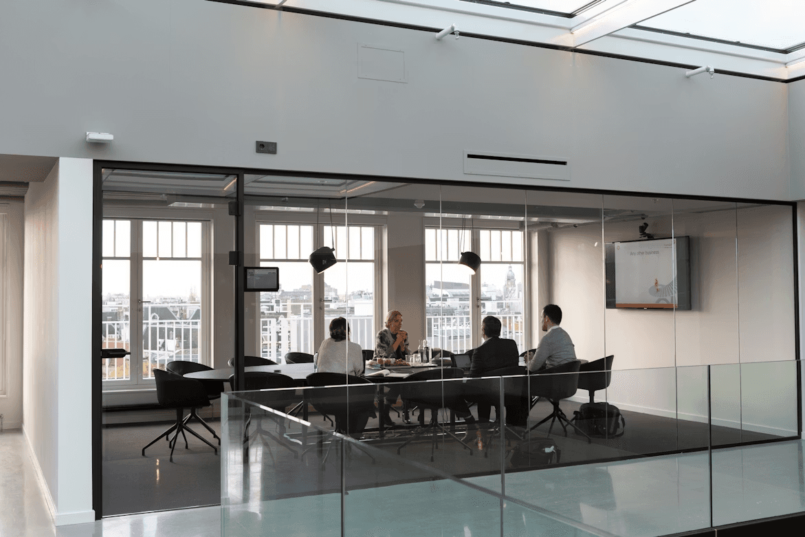A small group meeting in a glass-walled conference room, with people seated around a table and a presentation screen on the wall.