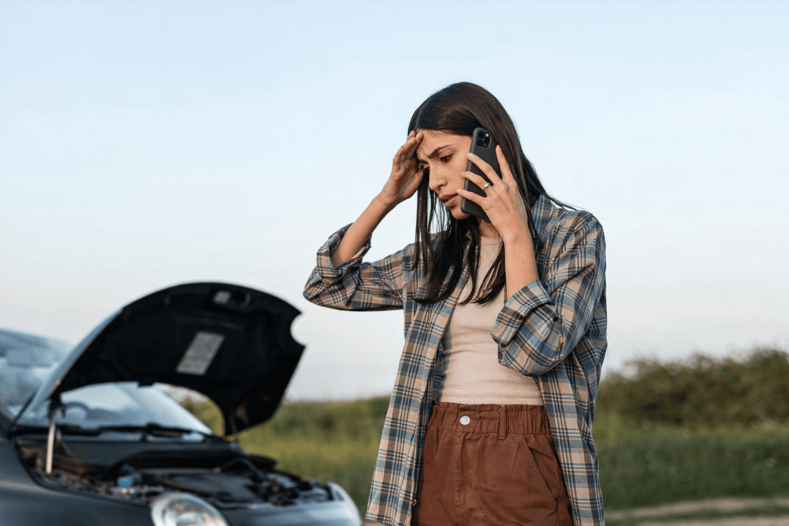 A worried woman on the phone stands beside a car with the hood up on a roadside, looking stressed as if she’s dealing with a breakdown.