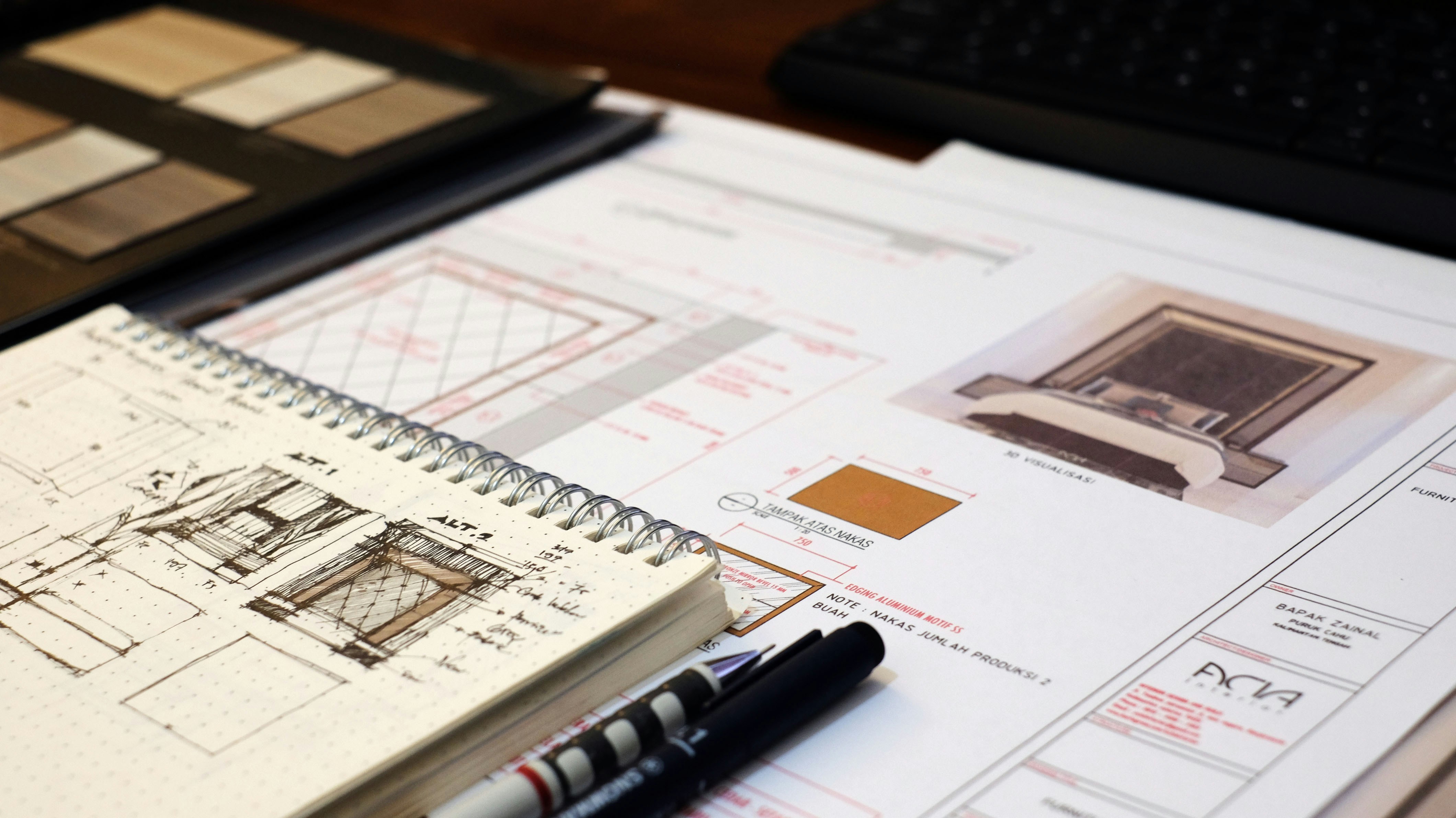 Architectural sketchbook on top of printed interior design plans with material samples and pens on a desk.