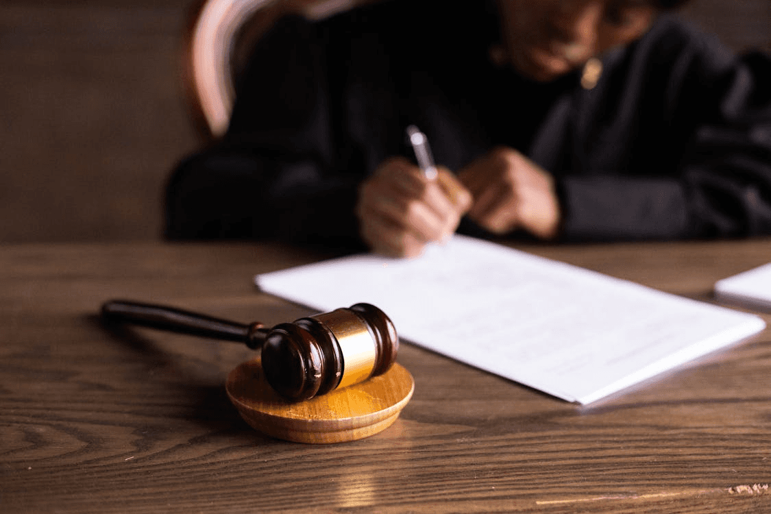 A judge’s gavel on a desk with a person signing paperwork in the background.