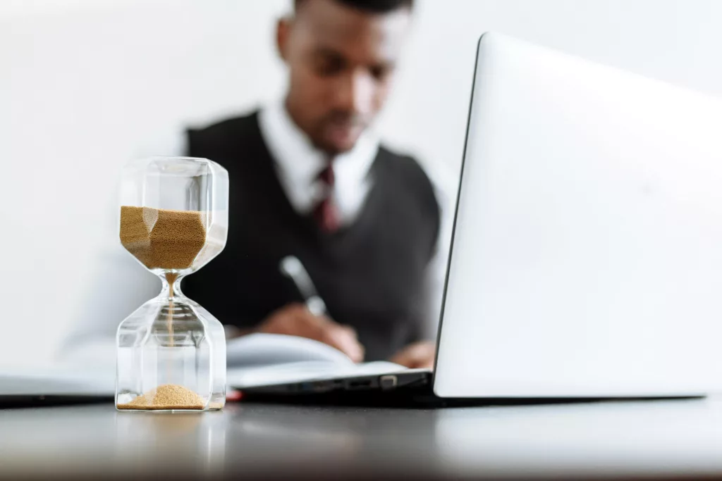 Hourglass on a desk with a person working on a laptop in the background