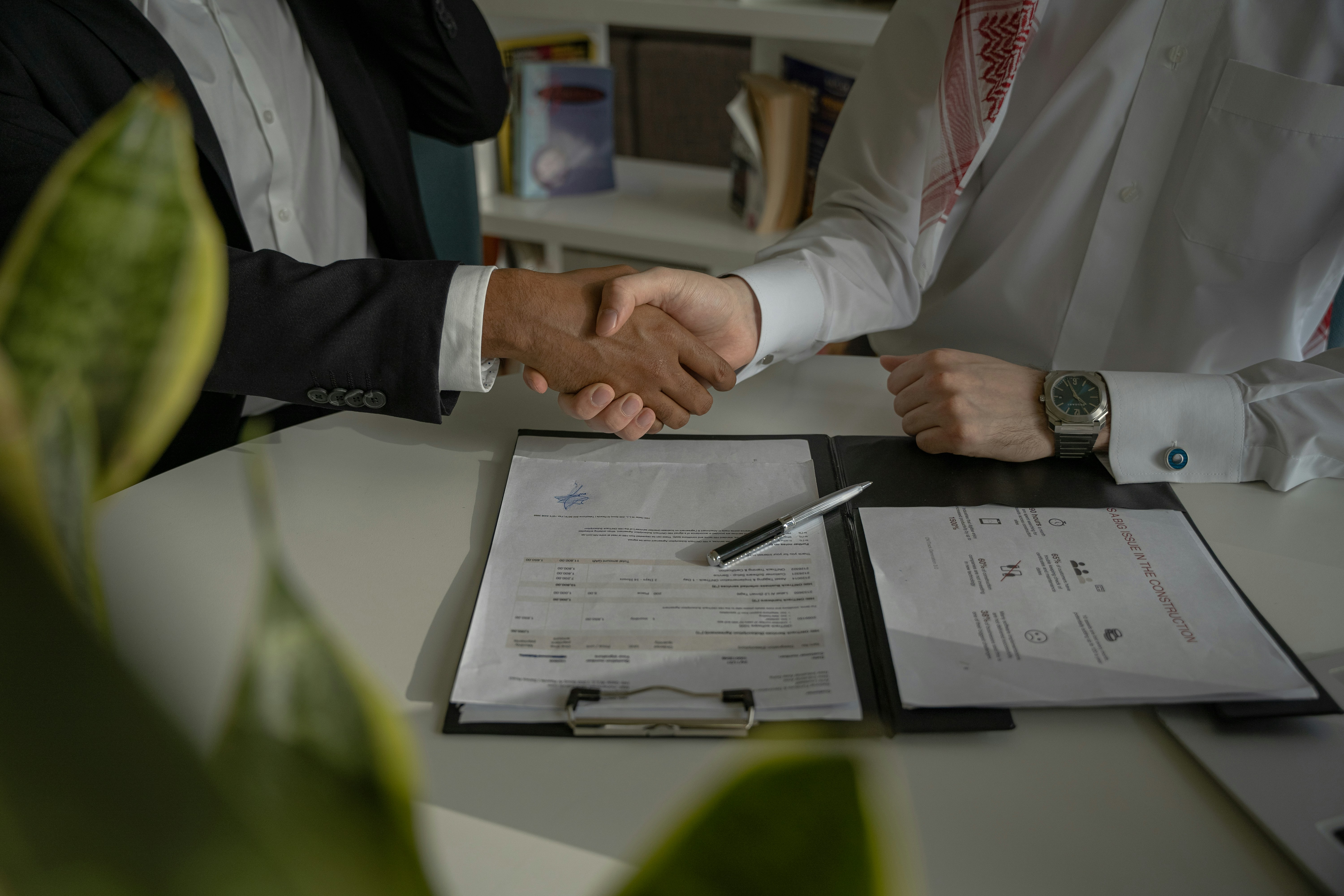 Two professionals shaking hands over signed documents and a pen on an office desk.