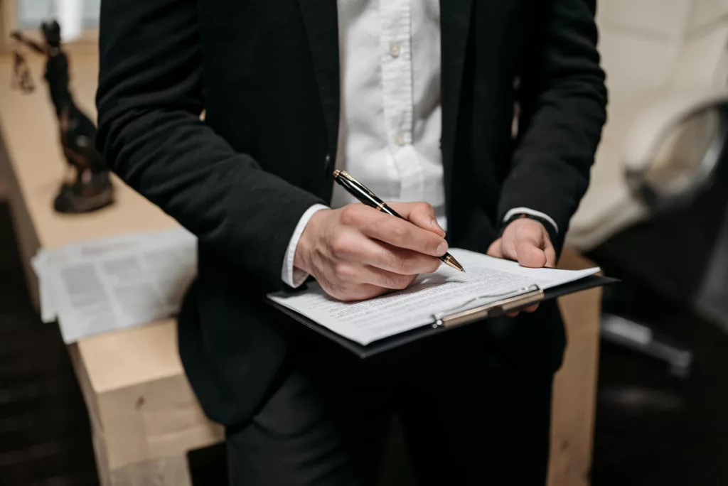 Person in formal attire writing on a clipboard near legal documents