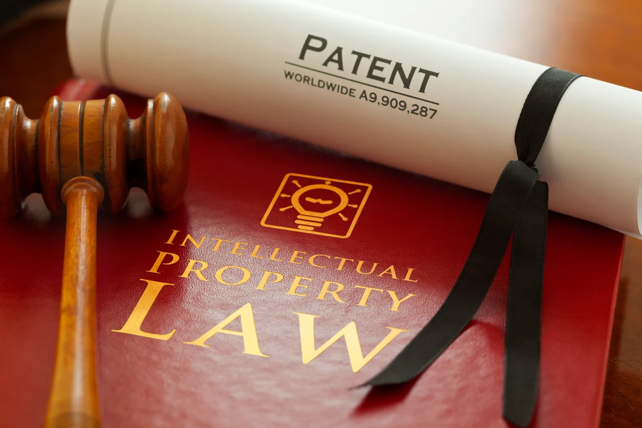 A close-up of a rolled PATENT certificate tied with a ribbon, resting on a red “Intellectual Property Law” book, with a judge’s gavel beside it. It’s a classic visual for protecting inventions and enforcing ownership rights through the legal system.