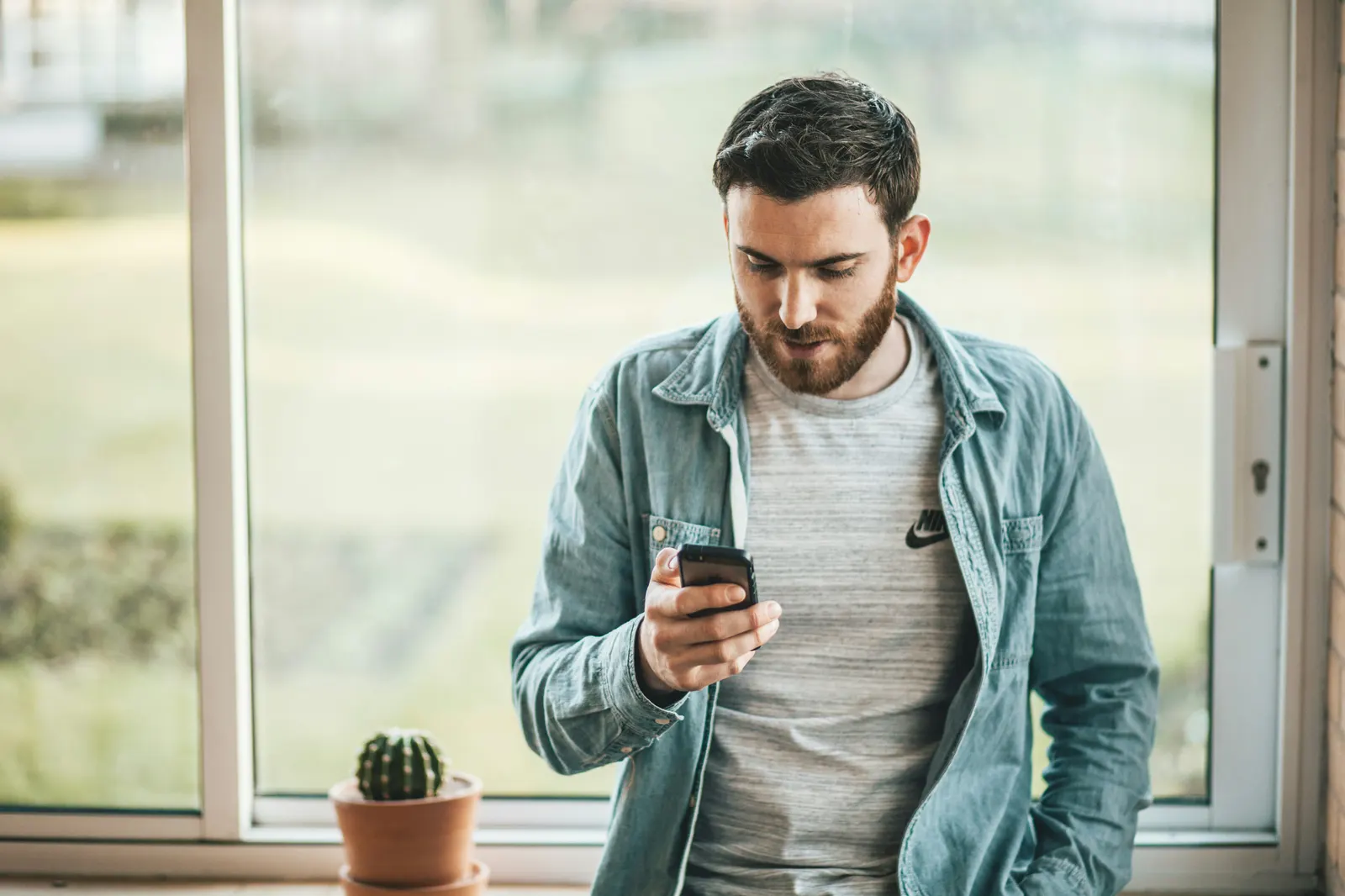 Bearded man standing by a window and looking at his smartphone.